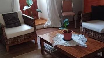Una habitación con dos sillas de mimbre, una mesa de centro de madera, una planta en maceta y vistas a través de una ventana con cortinas.