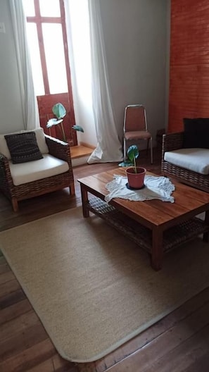 Una habitación con dos sillas de mimbre, una mesa de centro de madera, una planta en maceta y vistas a través de una ventana con cortinas.
