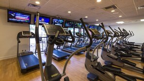 Un gimnasio con cintas de correr y una bicicleta estática, equipado con pantallas digitales y una pared de televisores que muestran canales de noticias.