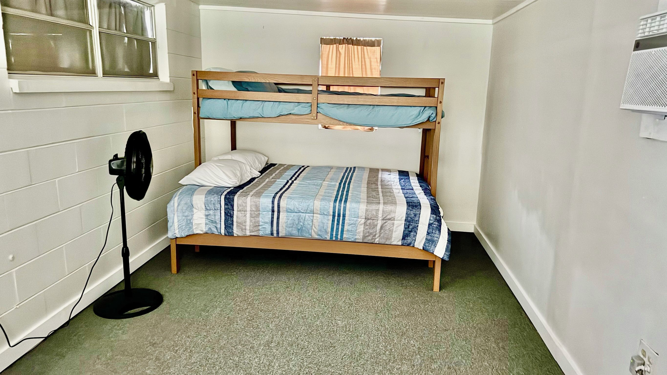 Uma cama beliche com colcha listrada, um ventilador e uma janela com cortinas.