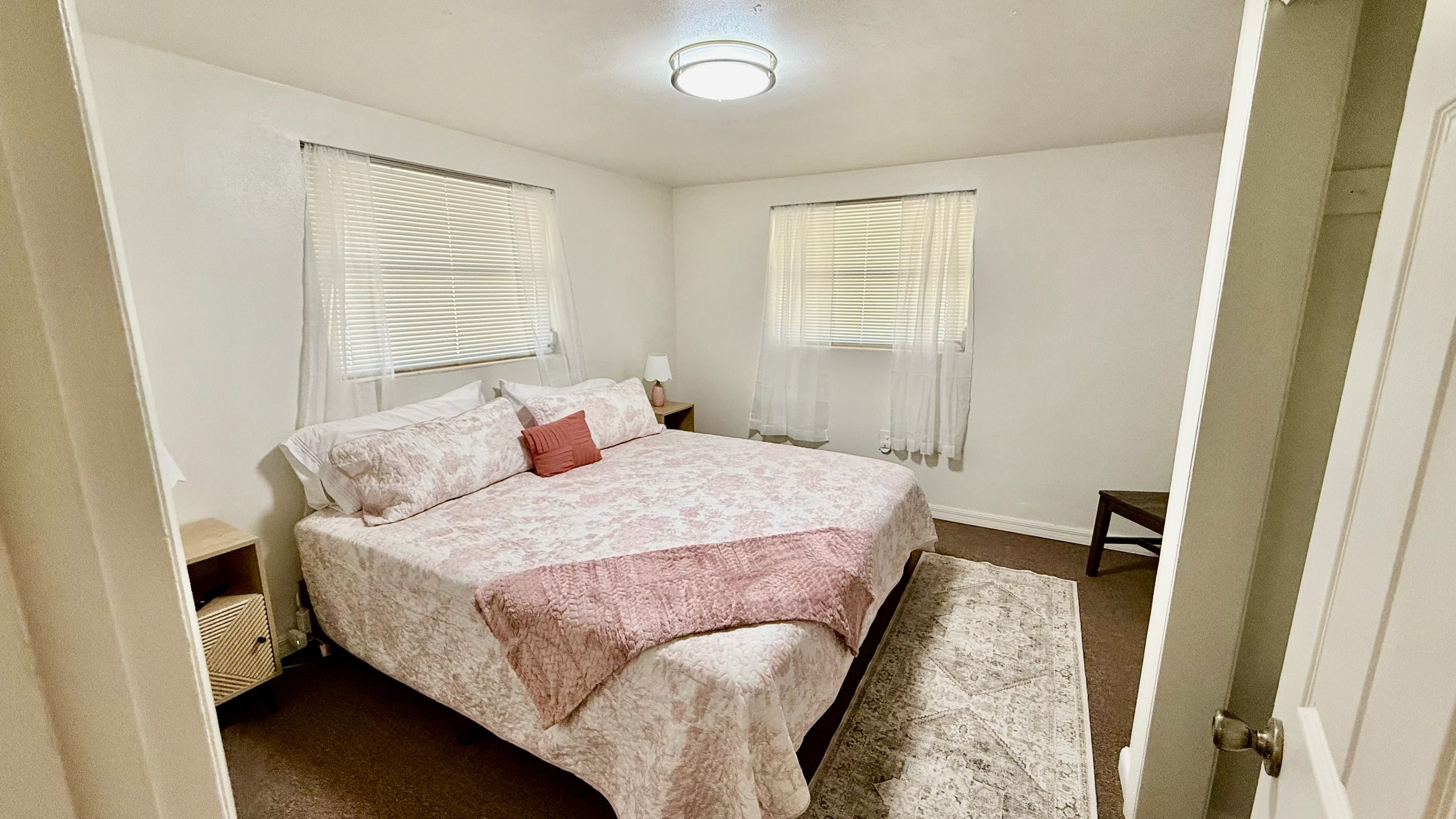 A bedroom with a bed, two bedside tables, a nightstand, a rug, and two windows with blinds.
