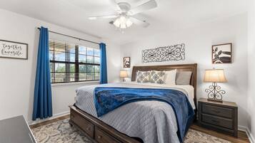 Un dormitorio con cama, mesitas de noche, ventilador de techo y ventana con cortinas.