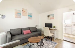 Ein modernes Wohnzimmer mit einem grauen Sofa, einem Holztisch, einem weißen Schreibtisch mit Computer und einem gemusterten Teppich.
