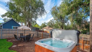 A backyard with a hot tub, a wooden deck, a table with chairs, and a blue building. - Golden Grove! Large Yard w/ Hot Tub Near Downtown (Colorado Springs)
