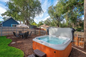 A backyard with a hot tub, a wooden deck, a table with chairs, and a blue building. - Golden Grove! Large Yard w/ Hot Tub Near Downtown (Colorado Springs)