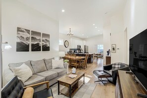 A modern living room with a sofa, coffee table, and a TV. There is a dining area with a table and chairs. The room has white walls and a chandelier.