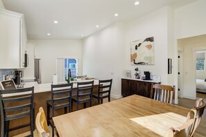 A modern kitchen with a dining area, featuring a wooden table and chairs, a coffee machine, and a large abstract painting on the wall.