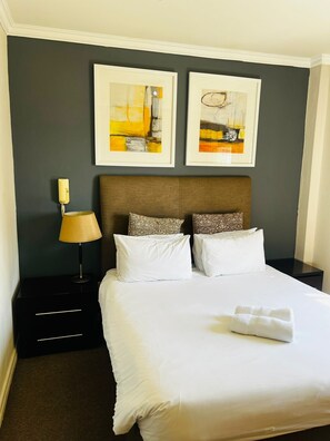 Habitación de hotel con una cama grande, dos almohadas, una lámpara de noche, una mesita de noche y dos cuadros enmarcados en la pared.