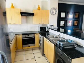 A compact kitchen with wooden cabinets, a microwave, a stove, and a washing machine. - Sandton Westpoint Luxury Apartments 191 (Sandton)