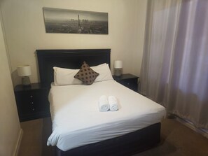 A hotel room with a neatly made bed, two bedside tables with lamps, a cityscape painting above the bed, and a window with curtains. - Sandton Westpoint Luxury Apartments 191 (Sandton)