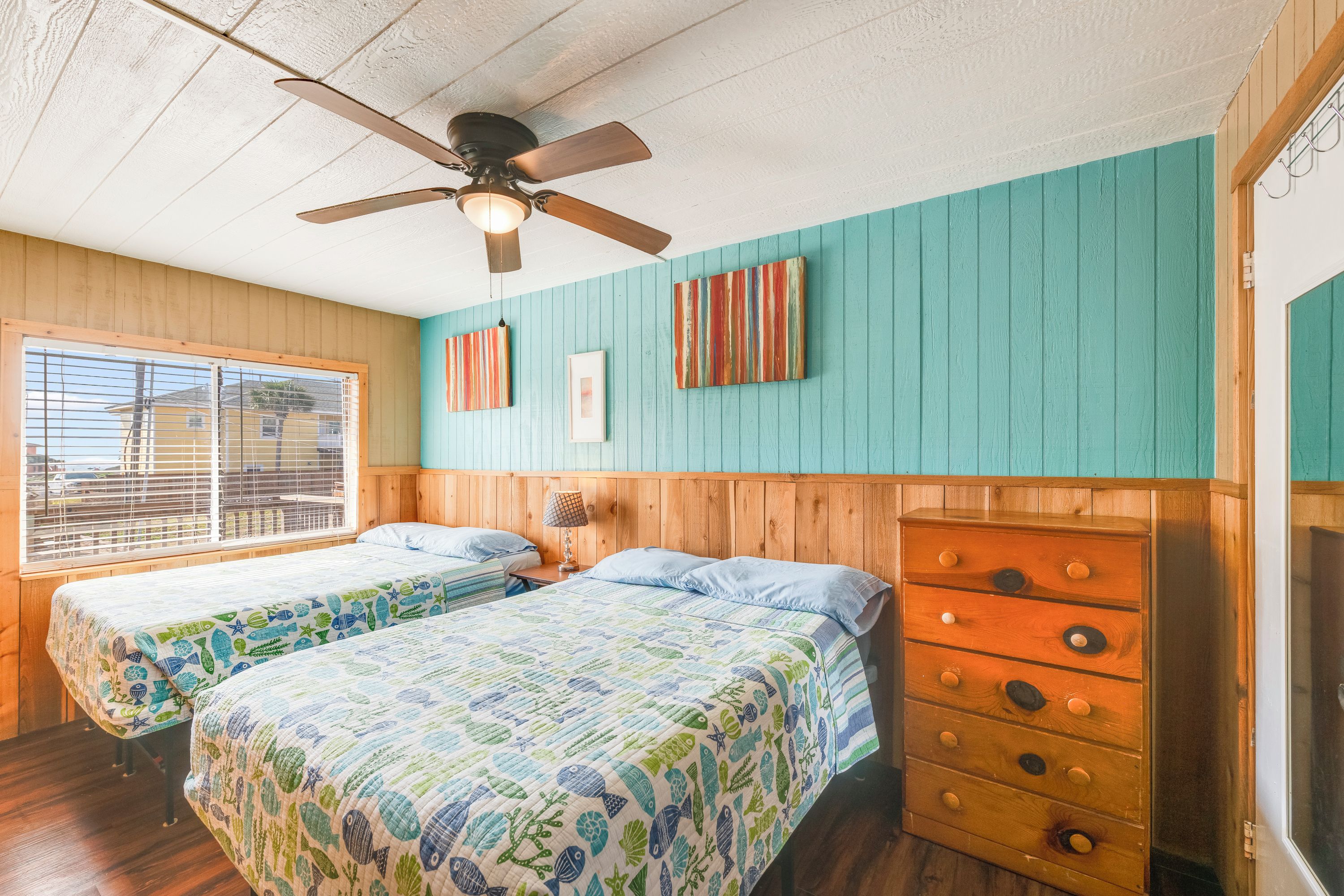 Une chambre avec deux lits, un ventilateur de plafond, une commode en bois et une fenêtre avec des stores.
