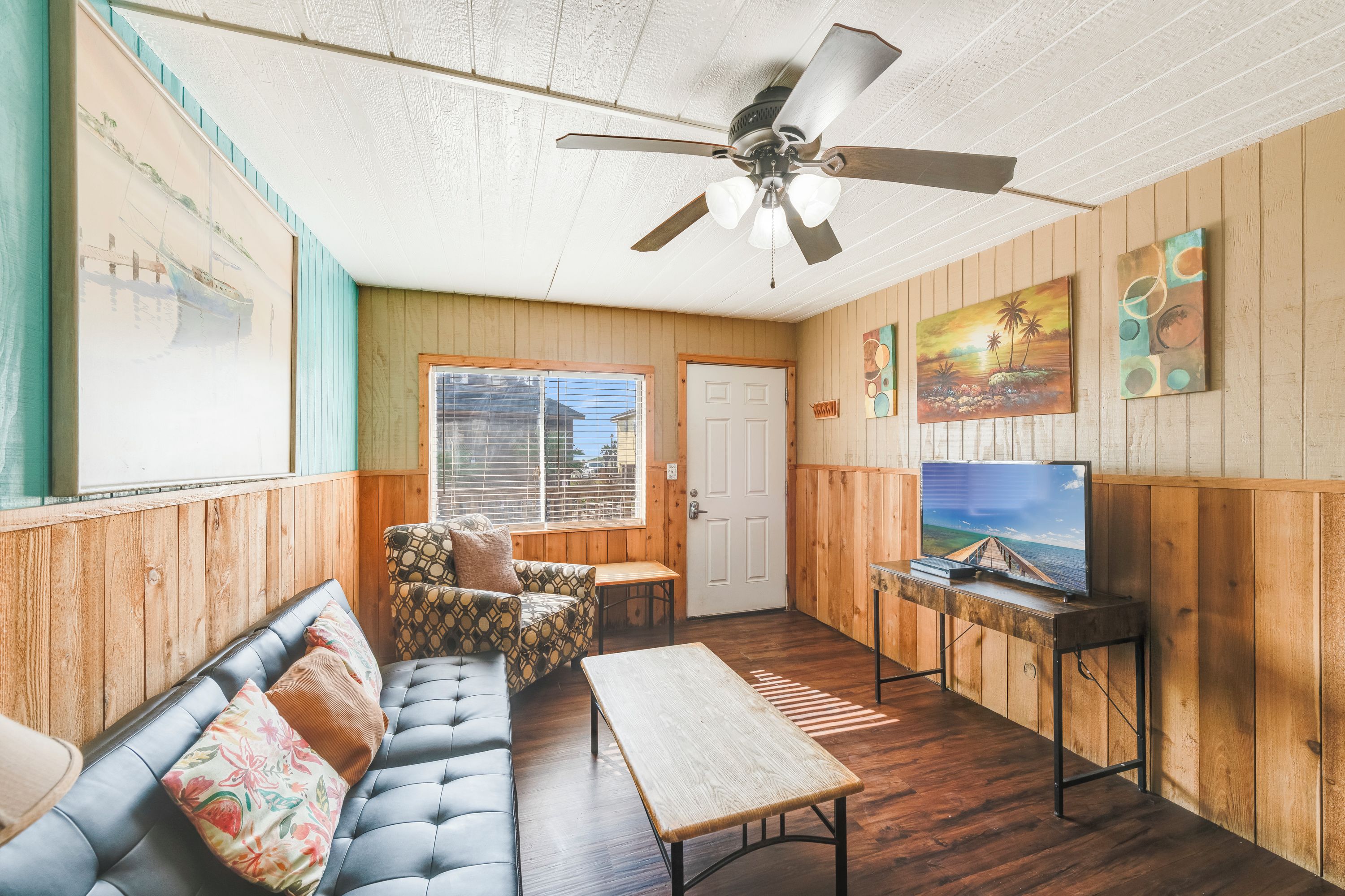 Une pièce dotée d’un ventilateur de plafond, d’un canapé, d’une table basse, d’un téléviseur posé sur un meuble en bois et de murs recouverts de panneaux de bois.