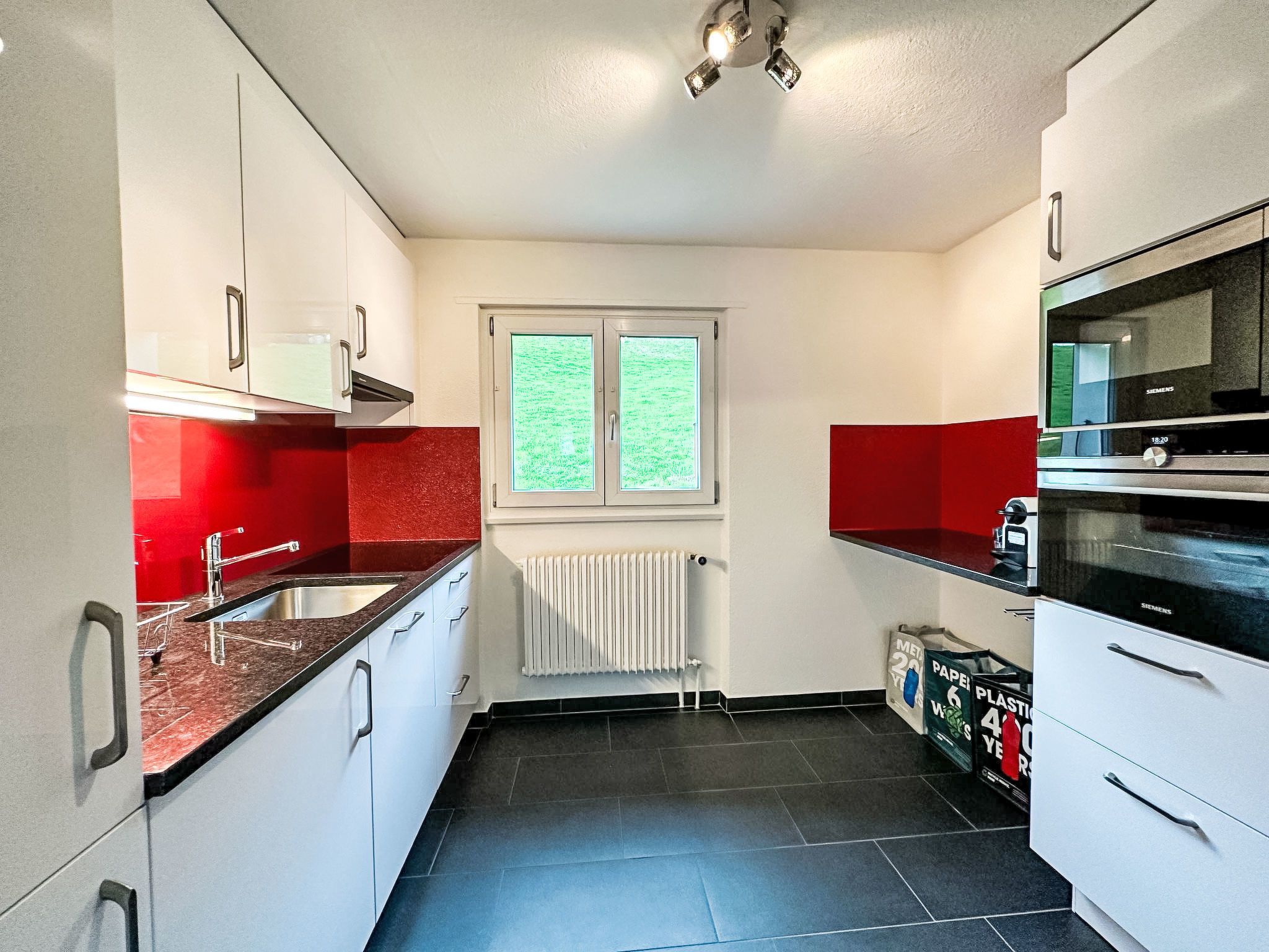 Una cocina con armarios blancos, encimera roja, horno Siemens y ventana con vistas.