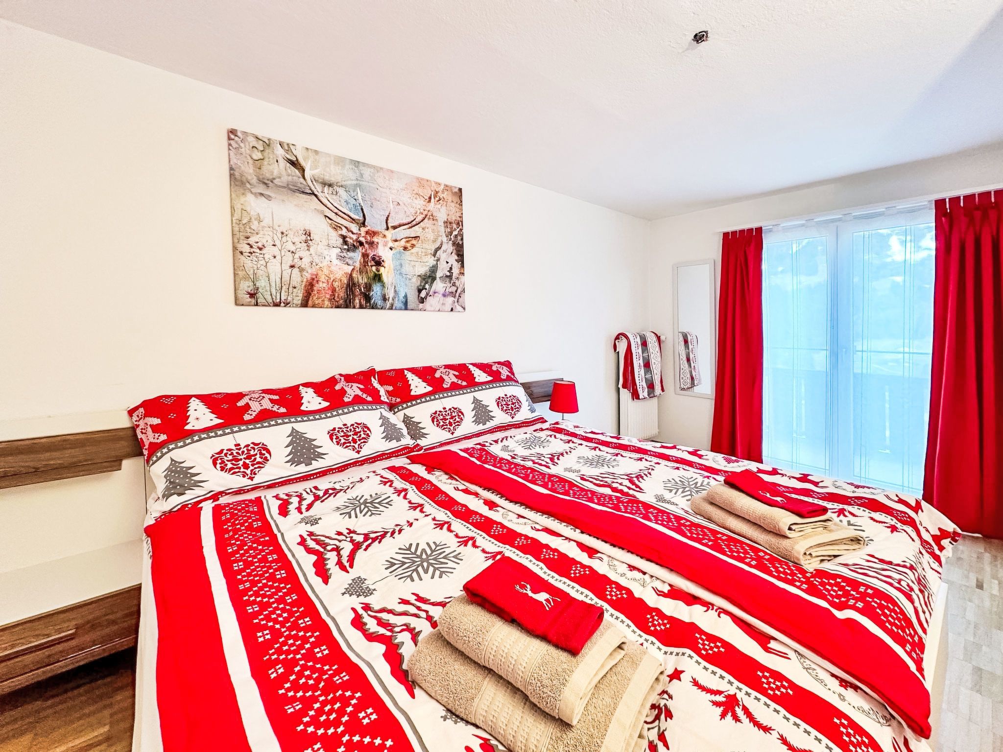 A hotel room with a large bed, a painting on the wall, and a window with red curtains.