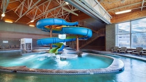 Ein großer Indoor-Wasserpark mit einer mehrstöckigen, blauen Rutsche, einem Schwimmbecken und einer Treppe, die zu einer höheren Ebene führt.