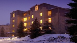 Un edificio de varios pisos con ventanas iluminadas, rodeado de nieve en el suelo y entre los árboles.