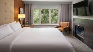 Ein Hotelzimmer mit einem großen Bett, einem Sessel, einem Fernseher, einem Kamin und einem Fenster mit Blick auf Bäume.