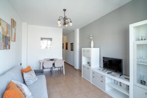 A modern living room with a grey sofa, a dining area with a table and chairs, a TV on a white cabinet, and a chandelier. - Apartment in Nerja Near Torrecilla Beach (Nerja)