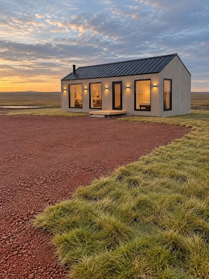 A modern, single-story cabin with large windows and a flat roof, situated in a natural landscape with a gravel driveway and grassy area.