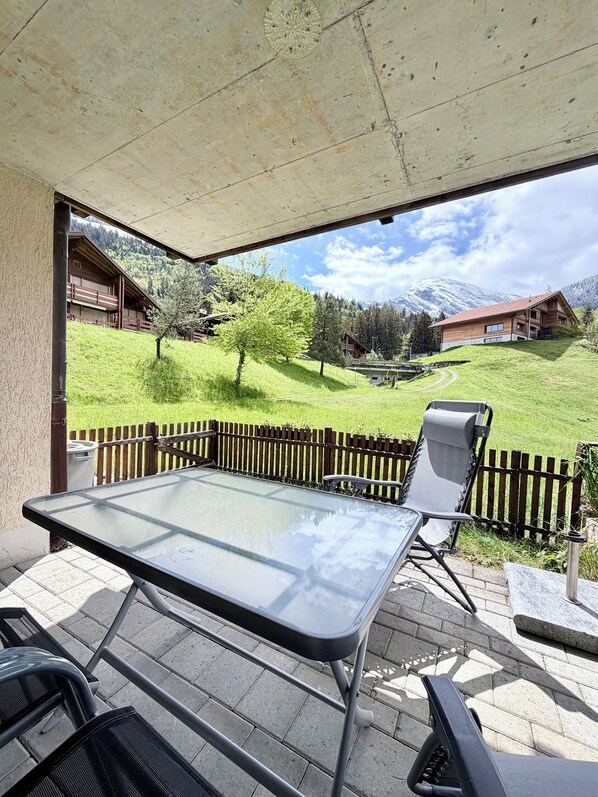 En udendørs terrasse med et bord og stole, et træhegn og udsigt til et bjerg og en bygning i baggrunden.