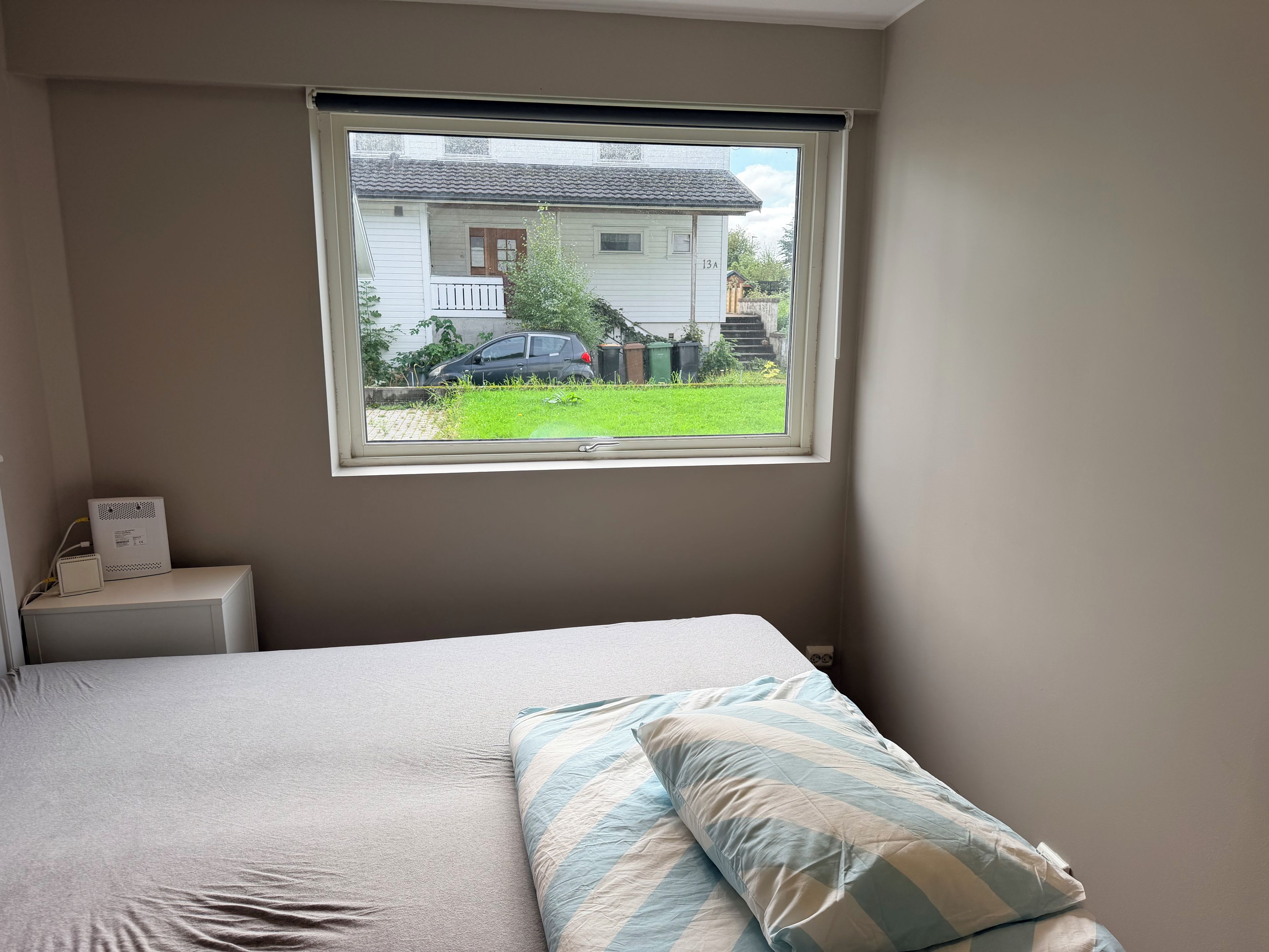 A bedroom with a bed, a nightstand, a window with a view of a house and car outside.