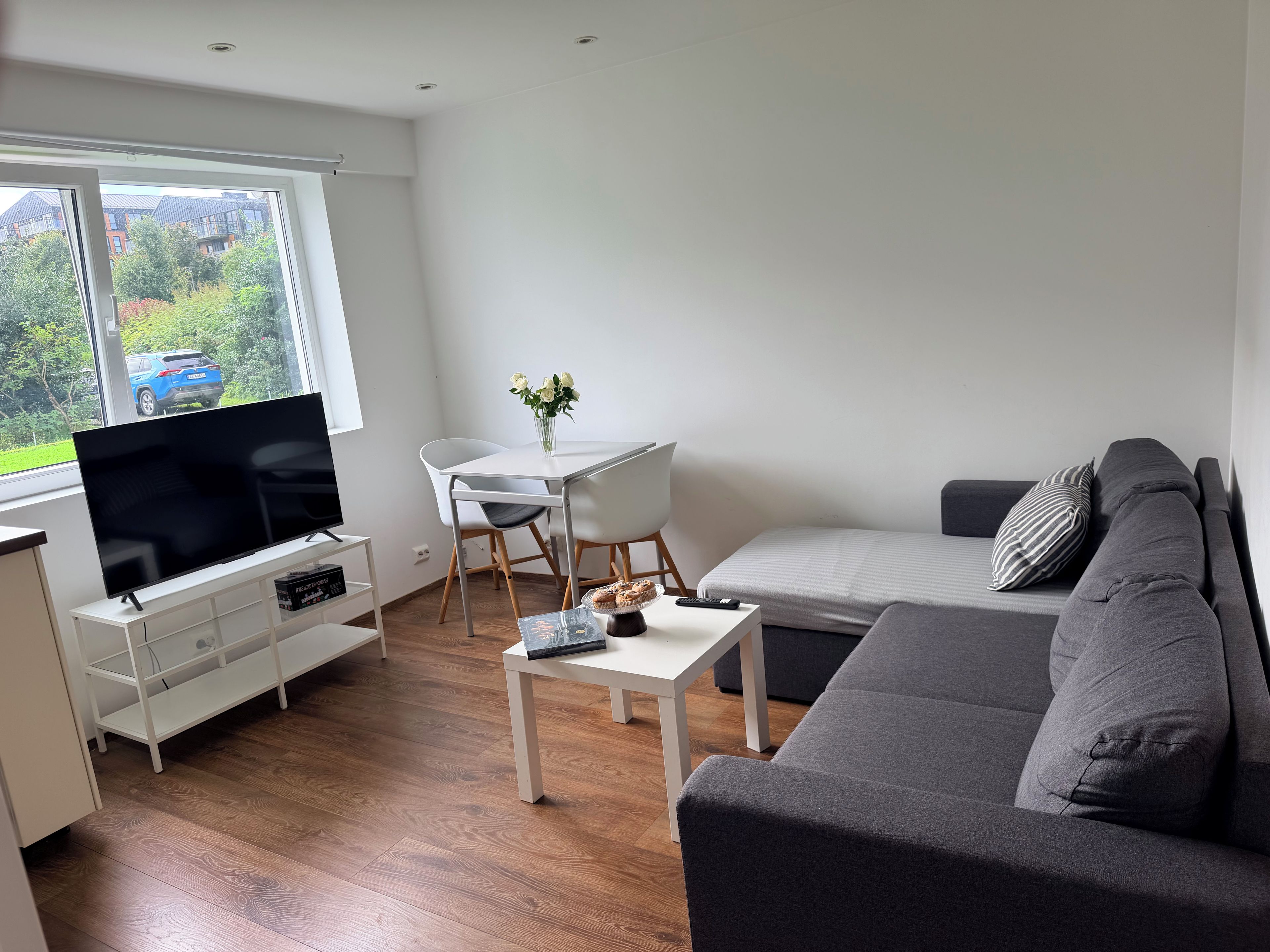 Ein modernes Wohnzimmer mit einem Flachbildfernseher, einem grauen Sofa, einem weißen Couchtisch und einem Essbereich mit weißem Tisch und Stühlen.