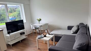 Ein modernes Wohnzimmer mit einem Flachbildfernseher, einem grauen Sofa, einem weißen Couchtisch und einem Essbereich mit weißem Tisch und Stühlen.