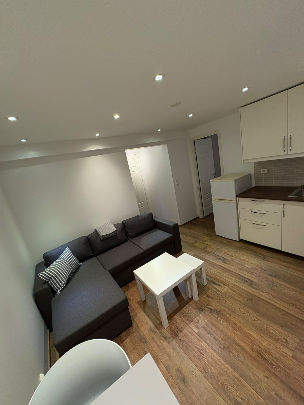Un espai de vida modern amb un sofà modular, una taula petita blanca i una zona de cuina compacta.