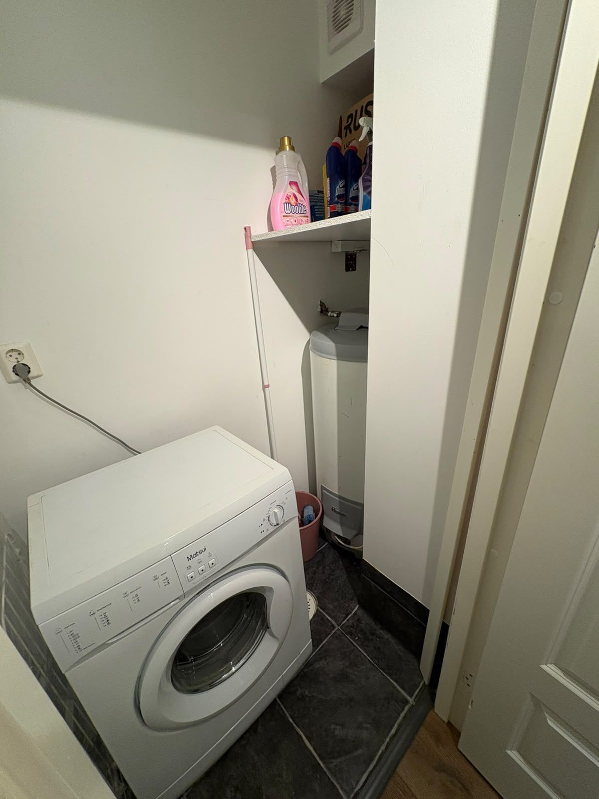 A compact laundry area with a front-loading washing machine, a shelf with cleaning supplies, and a small storage cabinet.