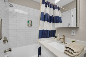 Casa de banho com banheira branca, lavatório com torneira dourada, cortina de chuveiro listrada em azul e branco e parede revestida a azulejos brancos.