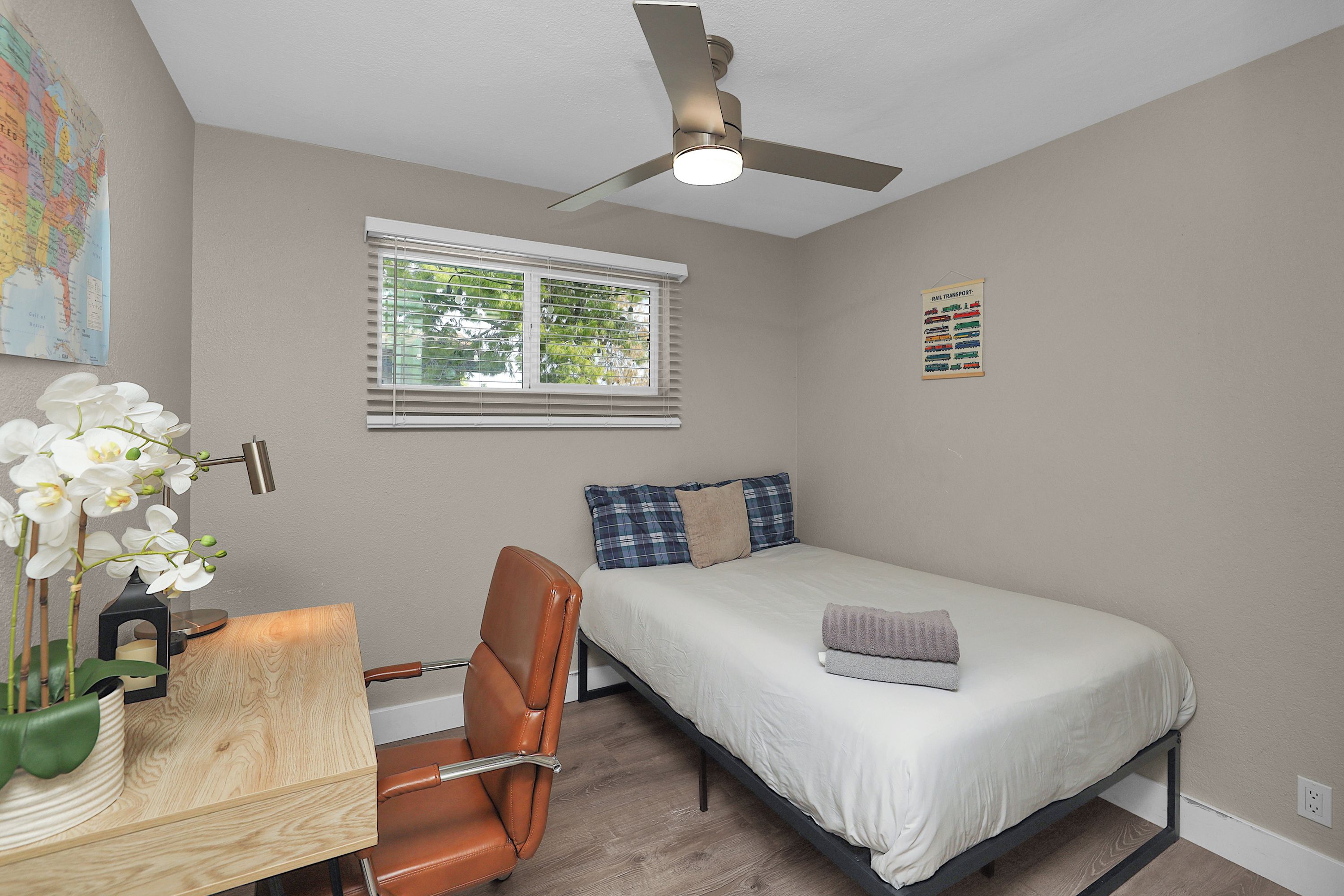 A bedroom with a bed, a desk with a chair, a ceiling fan, a window with blinds, and a wall map.