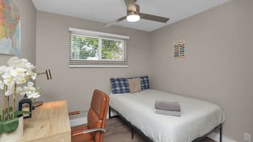 A bedroom with a bed, a desk with a chair, a ceiling fan, a window with blinds, and a wall map.