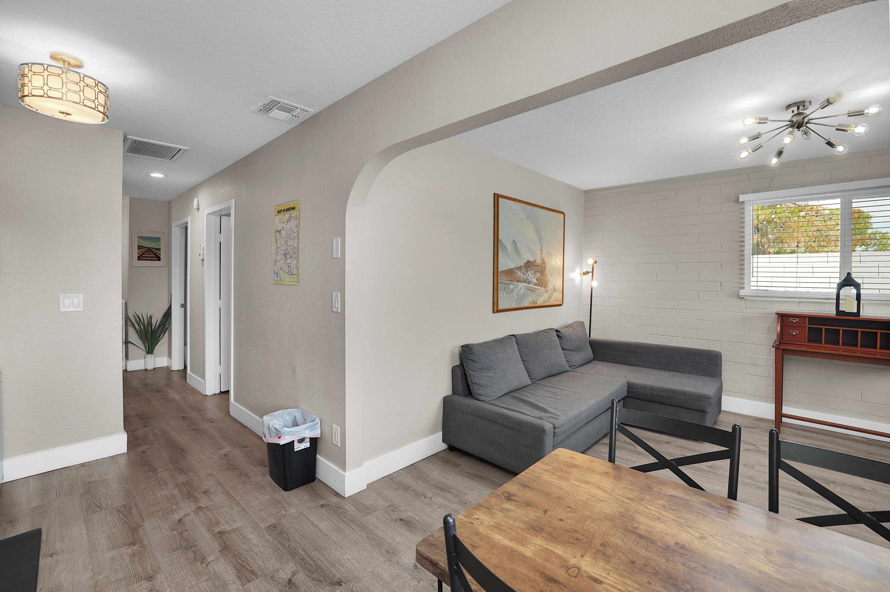 A modern living room with a gray sectional sofa, a wooden dining table, and a kitchen area with a brick backsplash.