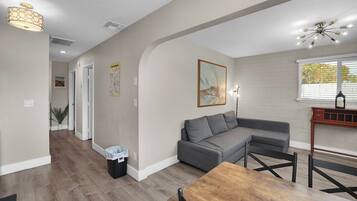 A modern living room with a gray sectional sofa, a wooden dining table, and a kitchen area with a brick backsplash.