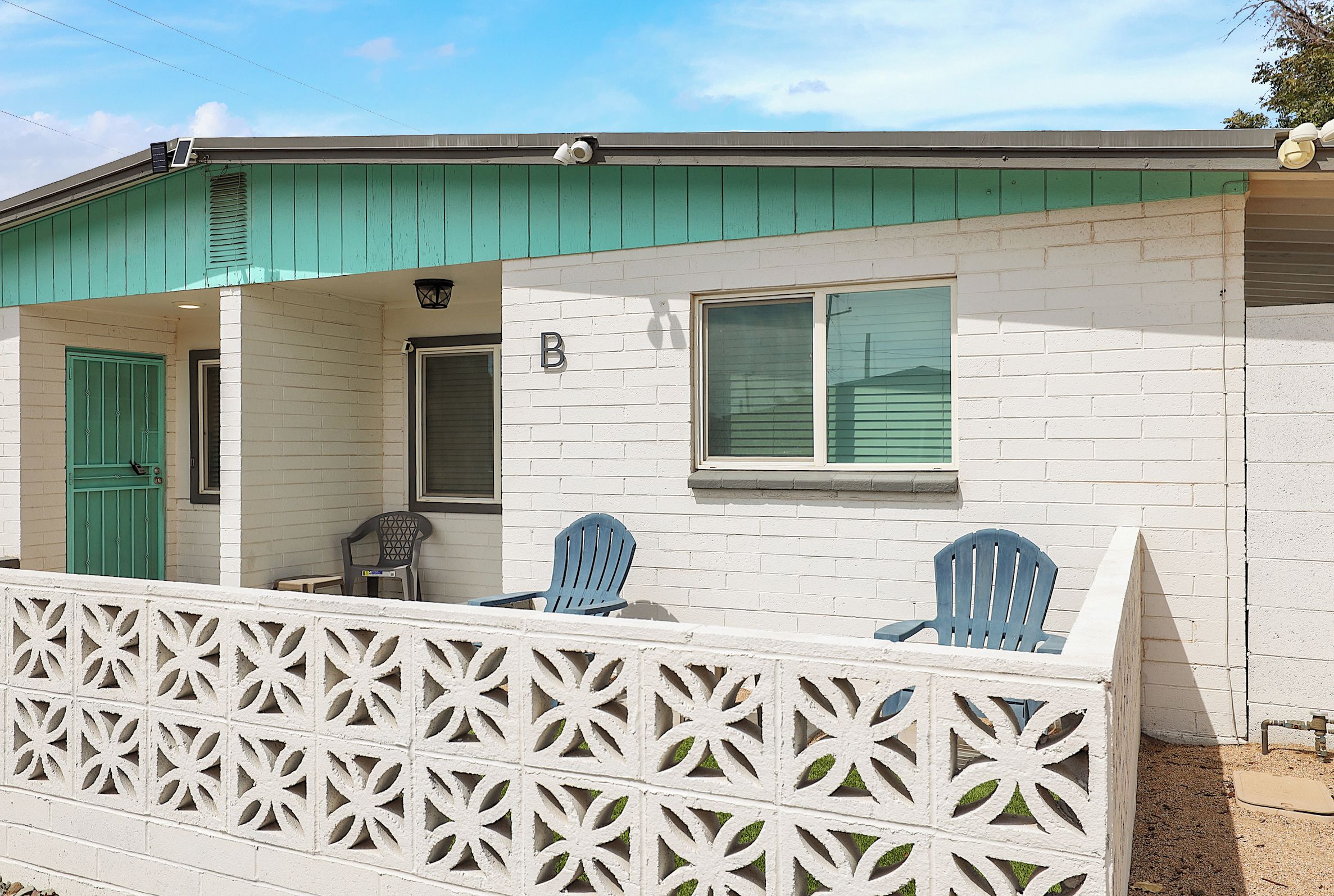 A small house with a green roof, a white fence with a geometric pattern, and two blue chairs outside.