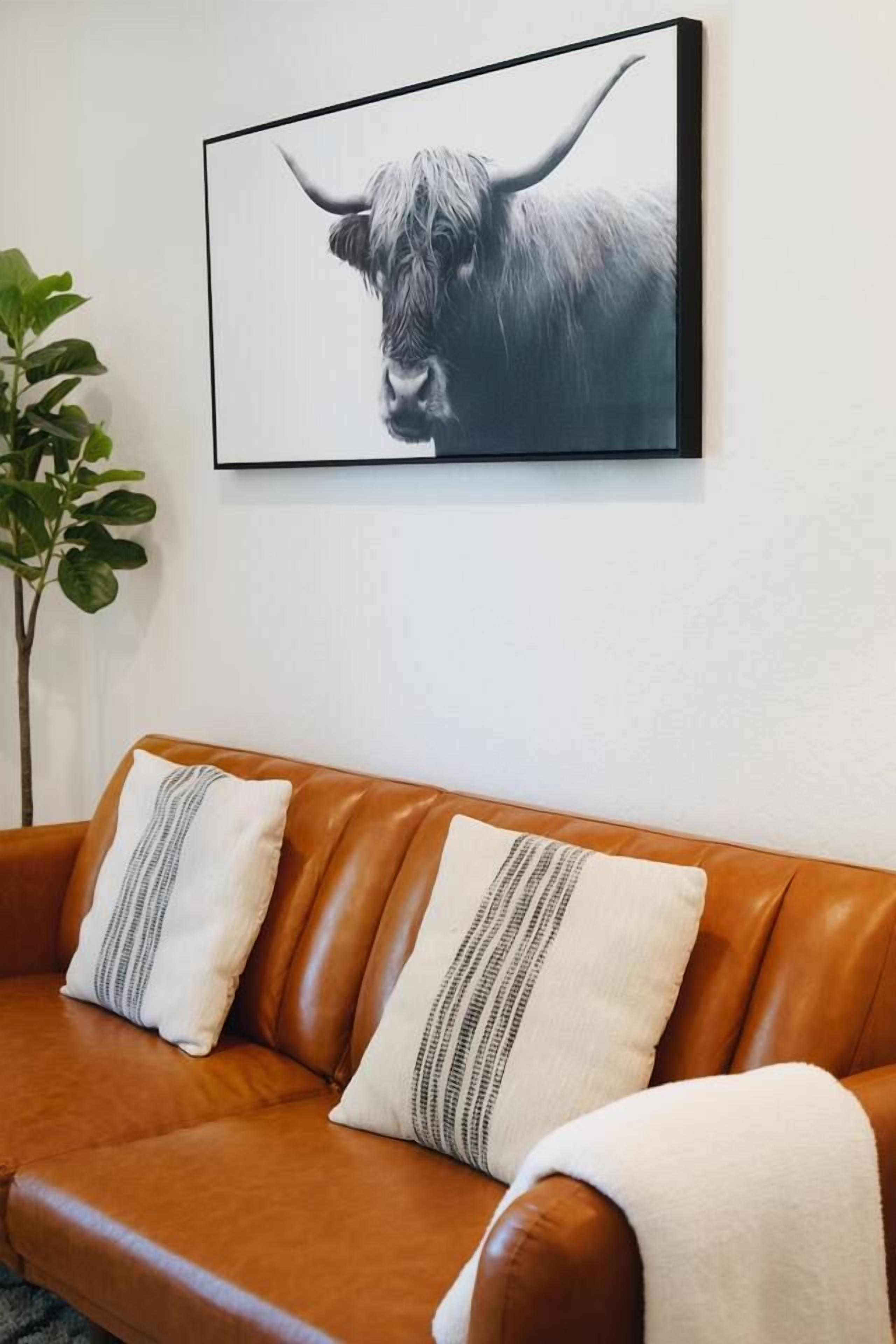 A brown leather sofa with two pillows, a framed picture of a bull on the wall, and a green plant to the left.