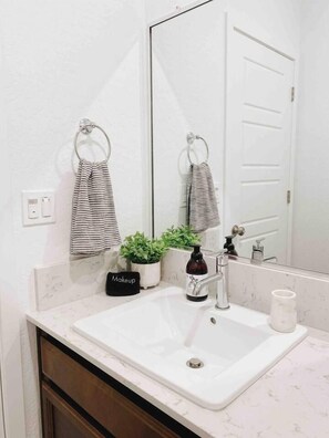 Un baño con un lavamanos blanco, un espejo, un perchero con toallas a rayas, una planta en maceta y un neceser.