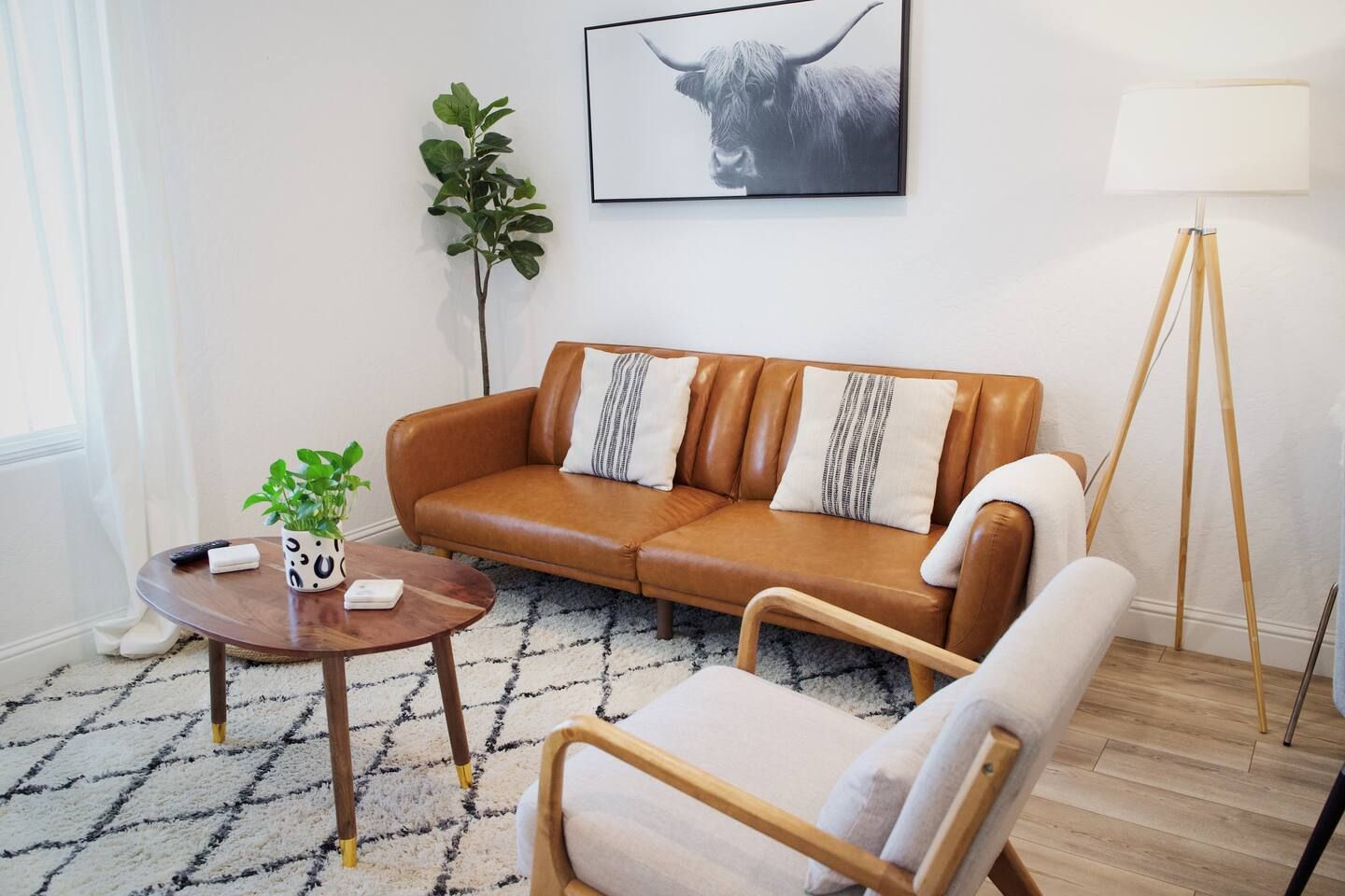 Una sala de estar con un sofá de cuero marrón, un sillón blanco, una mesa de centro de madera, una planta en maceta y una lámpara de pie.