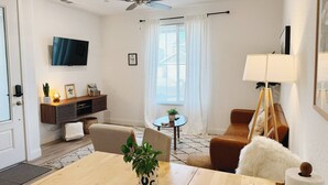 A modern living room with a wooden dining table, a sofa, a TV mounted on the wall, and a ceiling fan. - Cozy King Bed Suite | Near Shopping & DT Visalia (Visalia)