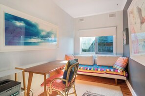Une pièce chaleureuse avec une table en bois, un fauteuil en osier, un canapé coloré et une grande fenêtre.