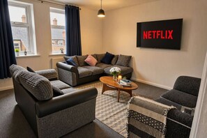 Ett vardagsrum med en soffgrupp, ett soffbord med böcker och ett ljus, och en väggmonterad tv som visar Netflix-logotypen.