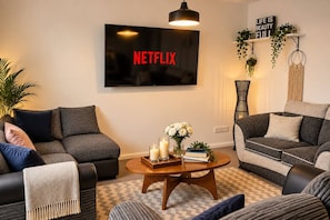 A living room with two sofas, a coffee table with books and candles, a TV mounted on the wall, and a shelf with plants and a motivational sign. - The Cozy Gateshead Escape (Tyne and Wear)