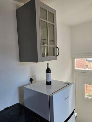 A kitchen with a white refrigerator, a wine bottle on top, a grey cabinet above, and a window with a view of a brick building. - The Cozy Gateshead Escape (Tyne and Wear)