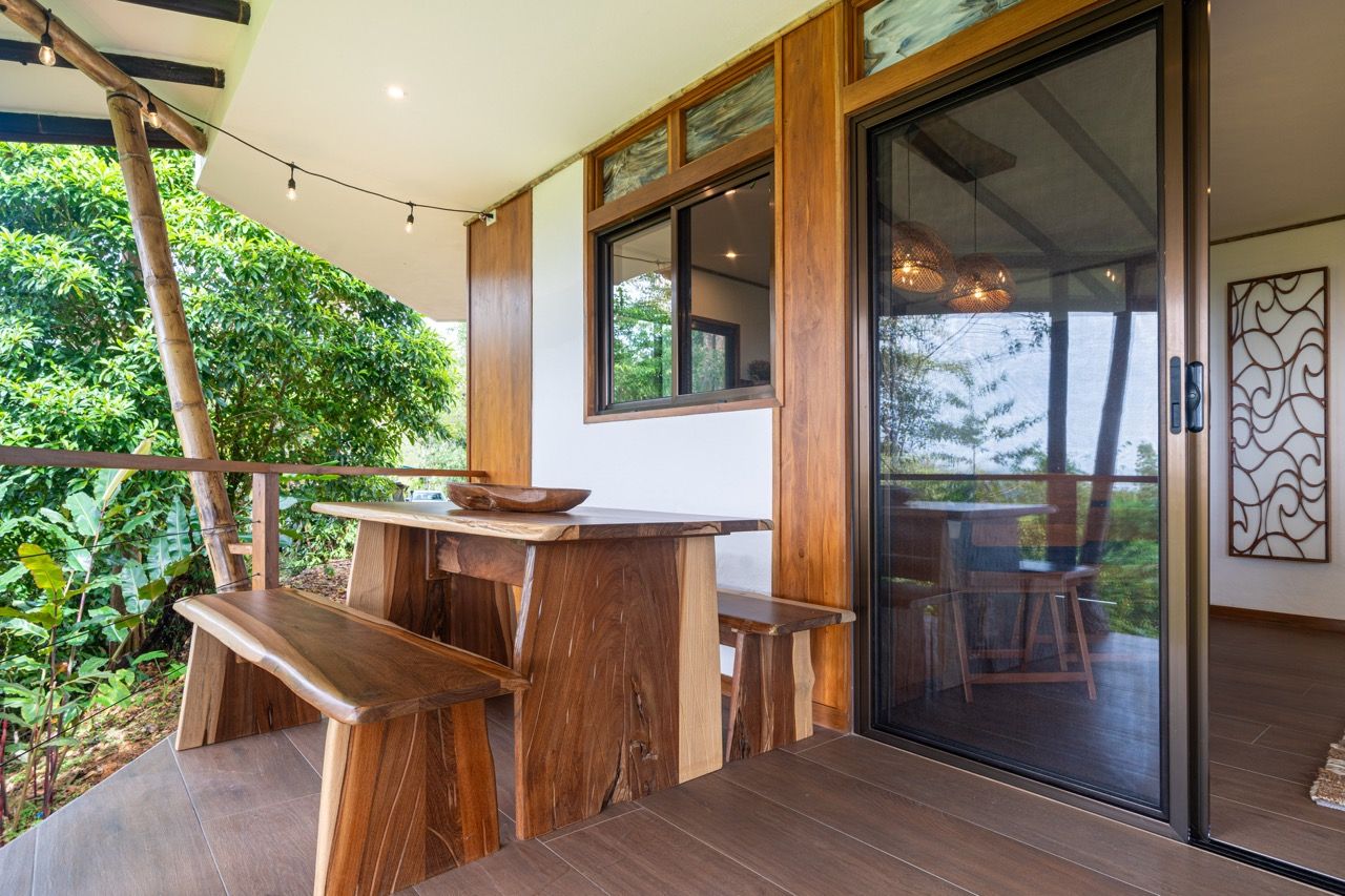 A wooden outdoor dining area with a table and benches, a glass door leading to a balcony, and a view of greenery.