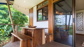 A wooden outdoor dining area with a table and benches, a glass door leading to a balcony, and a view of greenery.