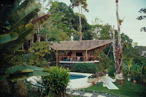 Uma casa isolada com telhado de palha, rodeada por vegetação exuberante e uma piscina.