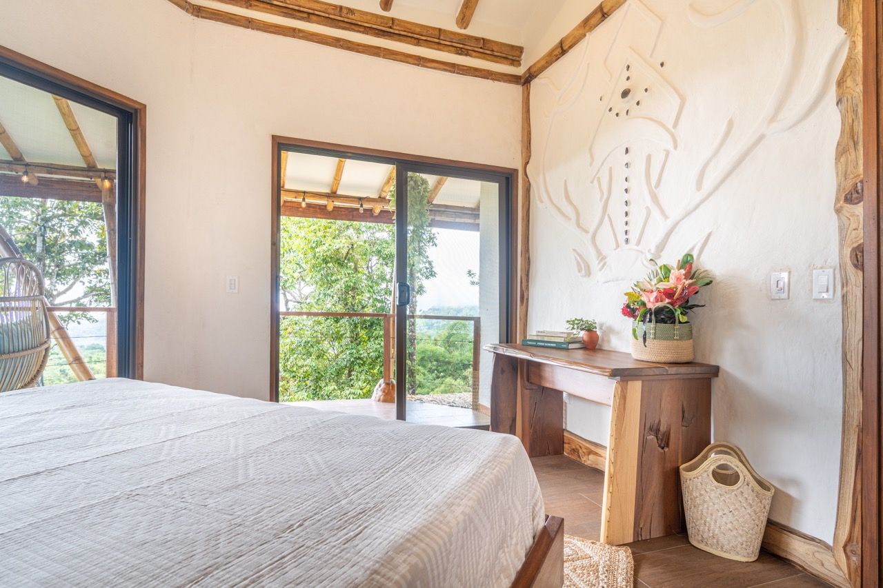 A bedroom with a large bed, a wooden desk, a chair, and a view of greenery through a sliding door.