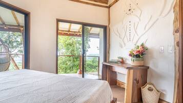 A bedroom with a large bed, a wooden desk, a chair, and a view of greenery through a sliding door.