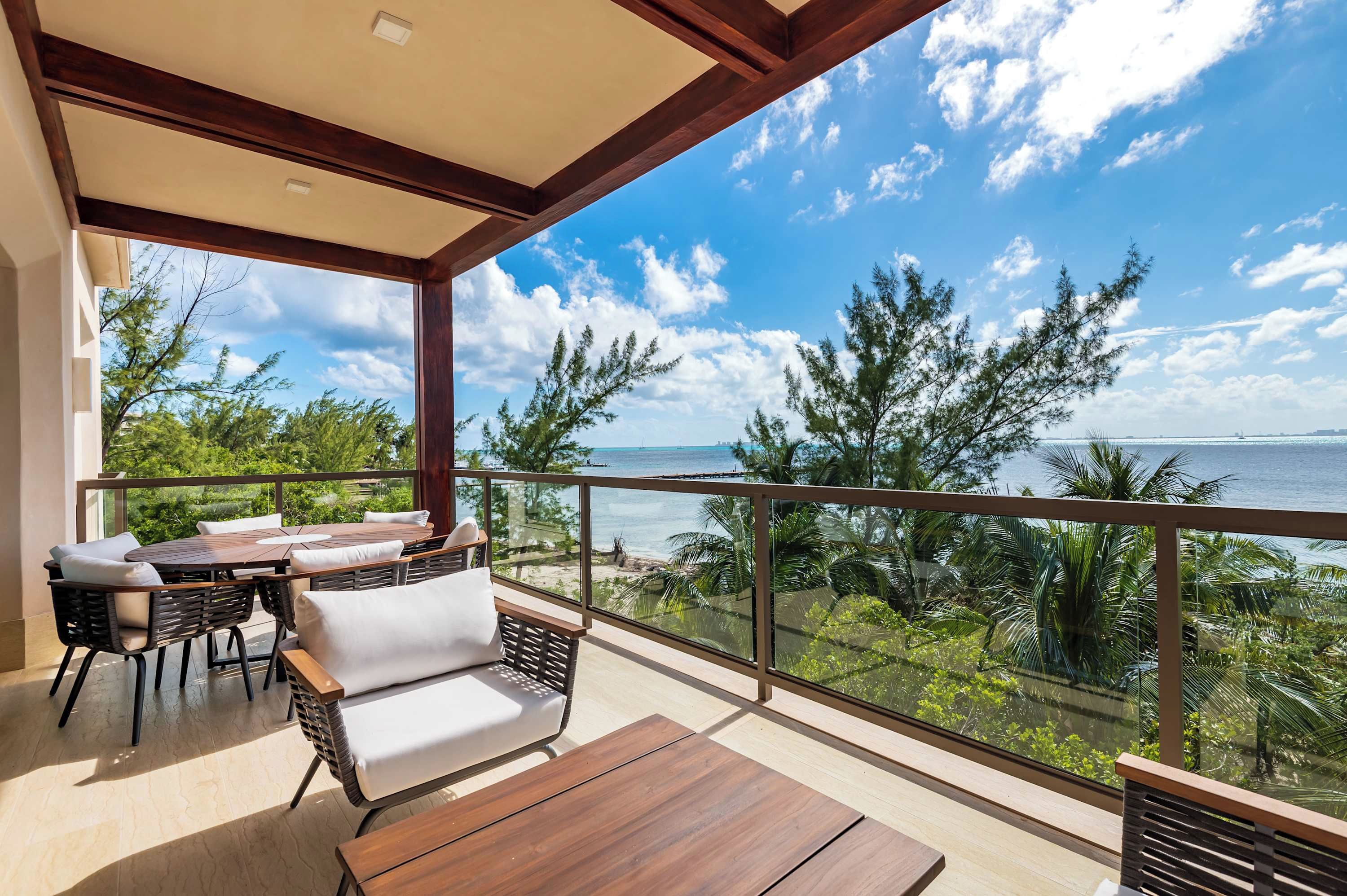 Un balcon doté d’une table et de chaises en bois, d’un canapé et d’une rambarde en verre, offrant une vue sur une plage et l’océan.