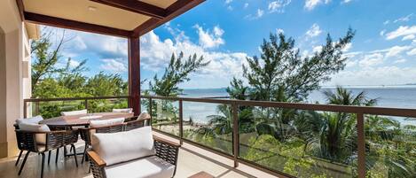 A balcony with a wooden table and chairs, a sofa, and a glass railing overlooking a beach and ocean.